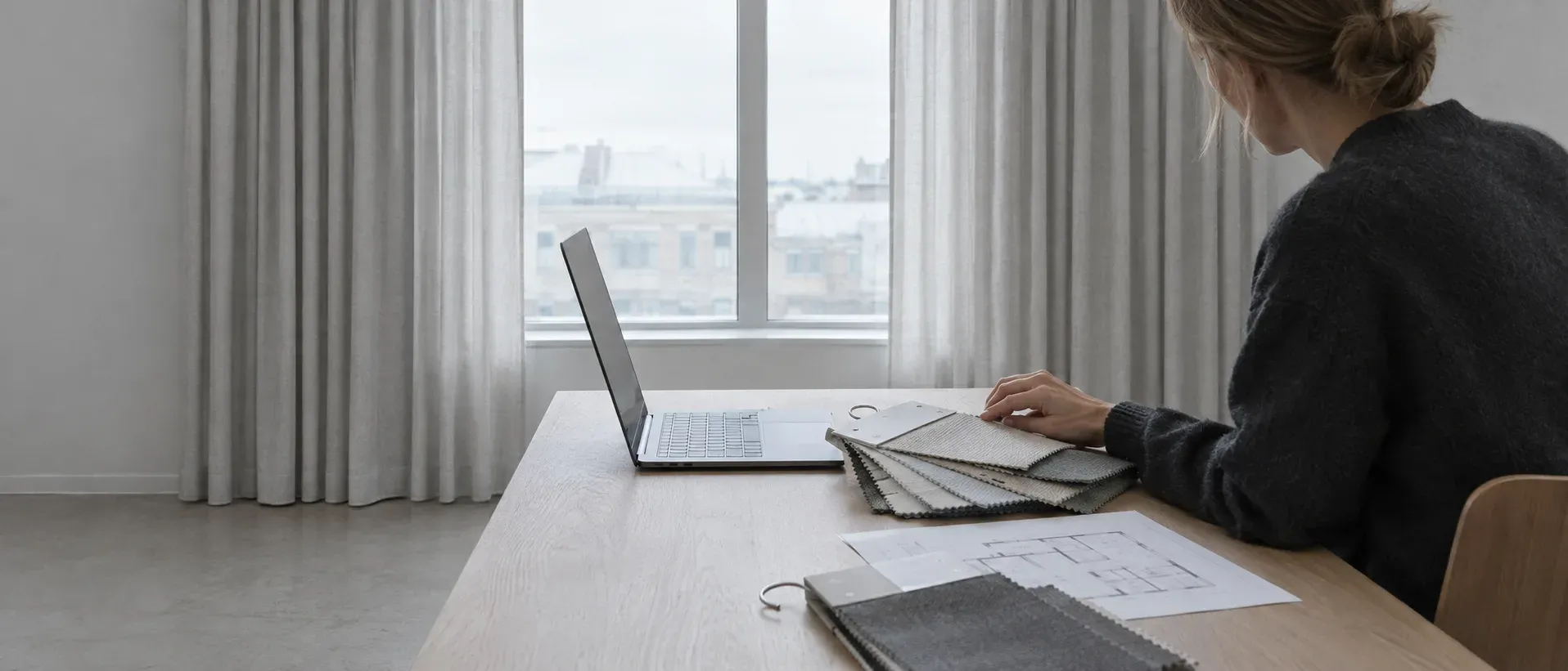 Skrivbord vid ett fönster med en dator, tygprover och en planritning
